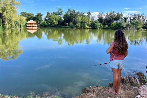 Una niña pescando junto a un lago tranquilo con árboles y una cabaña de madera al fondo en Francia.
