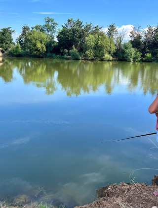 Une fille pêche au bord d’un lac paisible avec des arbres et un chalet en bois à Domaine des Feuilles.