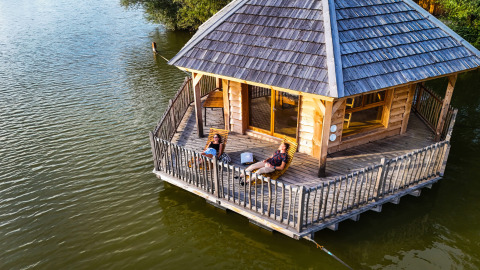 Twee personen genieten op het terras van cabine Ruche, een houten lodge op het water van een rustig meer.