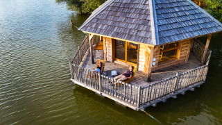 Due persone si rilassano sulla terrazza della cabine Ruche, un lodge in legno sull’acqua di un lago tranquillo.