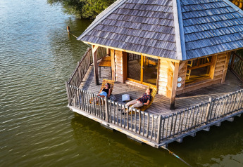 Zwei Personen entspannen auf der Terrasse von cabine Ruche, einer Holzlodge, die auf einem See schwimmt.