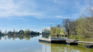 La cabina a specchio Cabine Spa Miroir riflette il paesaggio sereno del lago sotto un cielo azzurro.