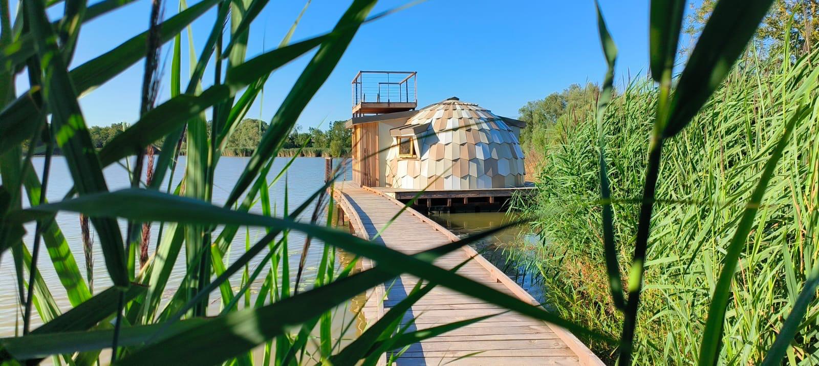 Houten pad leidt naar Cabine Spa Les Flots aan het water, omringd door riet en onder blauwe lucht.
