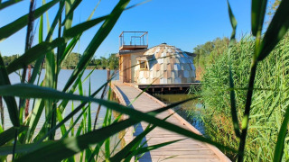 Passerella in legno verso Cabine Spa Les Flots sul lago, circondata da canne e cielo sereno.