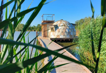 Pasarela de madera hacia la Cabine Spa Les Flots sobre el lago, rodeada de juncos y cielo azul.