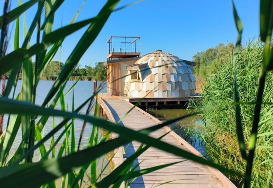 Houten wandelpad naar Cabine Spa Les Flots op het meer, omgeven door riet en een heldere lucht.