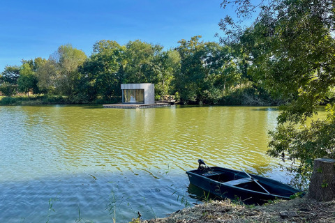 Moderne Lumipod Flottant-hut drijvend op een rustige plas, met groene bomen en boot op de voorgrond.