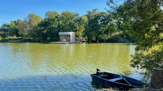 Moderna cabina Lumipod Flottant che galleggia su un lago tranquillo, con barca nera e alberi verdi intorno.