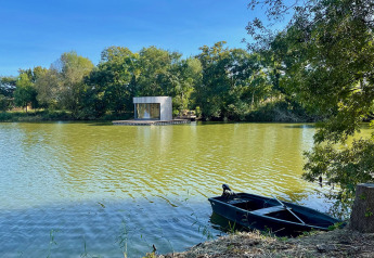 Moderna cabina Lumipod Flottant che galleggia su un lago tranquillo, con barca nera e alberi verdi intorno.