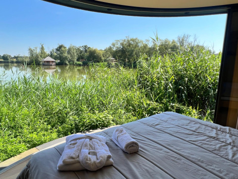 Uitzicht vanuit Lumipod île-hut met bed, groene natuur, een meer en een huisje in de verte.