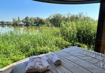 Zicht vanuit de Lumipod île-hut met een bed, groene oever, meer en een hutje in de verte.