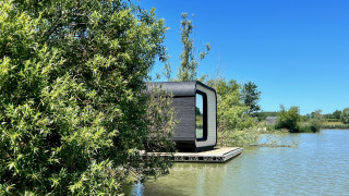 Moderna cabina Héron che galleggia su un lago tranquillo, parzialmente nascosta da alberi e natura.