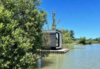 Eine moderne Héron-Hütte am See, teilweise von Bäumen verdeckt, bei klarem Himmel und Natur.