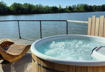 Jacuzzi exterior y tumbonas de madera en una terraza junto al lago en Cabine Spa Les Flots, Domaine des Feuilles.