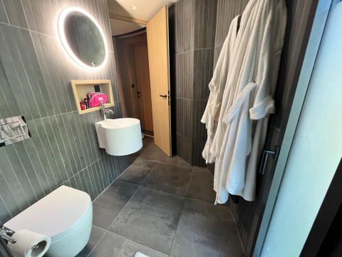 Moderne badkamer in een hut met grijze tegels, verlichte ronde spiegel en witte badjassen aan de muur.