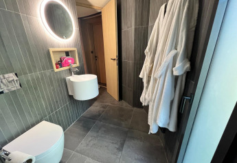 Bagno moderno in una baita con piastrelle grigie, specchio tondo illuminato e accappatoi bianchi appesi.