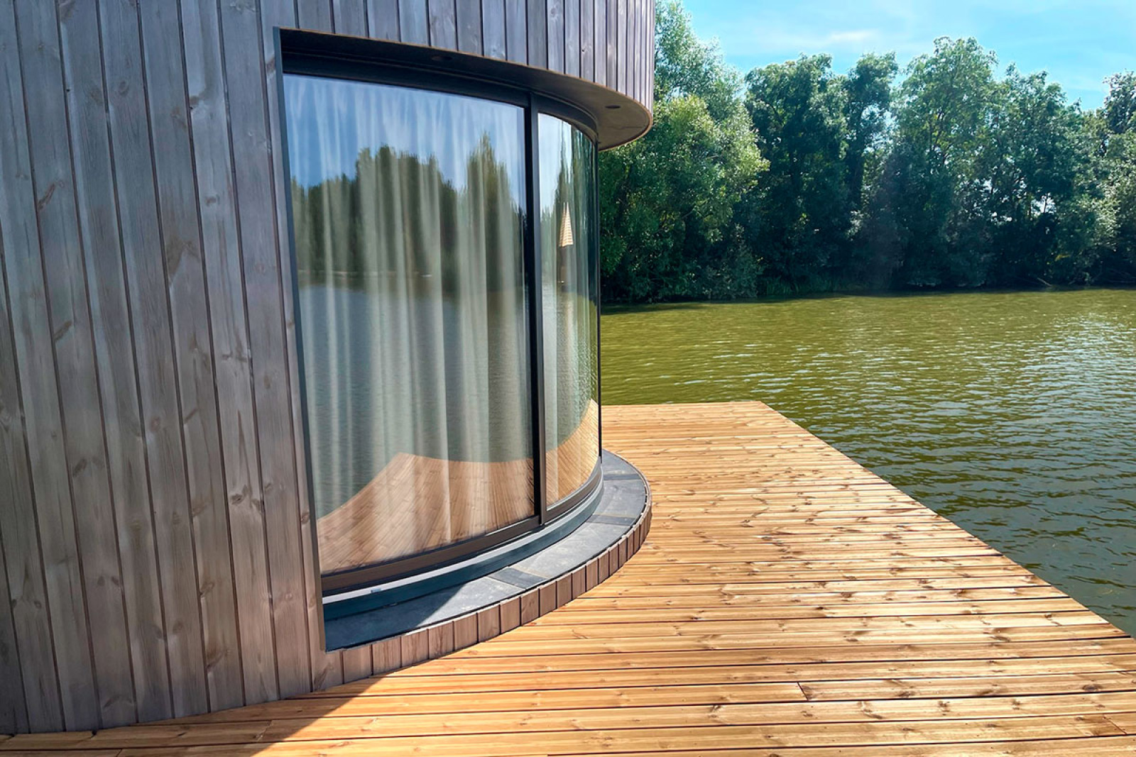 Gebogen glazen raam met gordijn in een hut aan het water, houten terras bij Lumipod Flottant, Frankrijk.