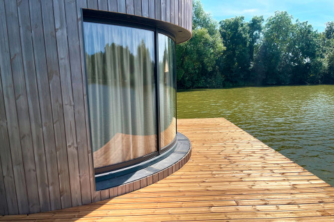 Gebogen glazen raam met gordijn in een hut aan het water, houten terras bij Lumipod Flottant, Frankrijk.