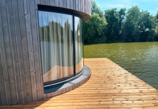Gebogen glazen raam met gordijn in een hut aan het water, houten terras bij Lumipod Flottant, Frankrijk.