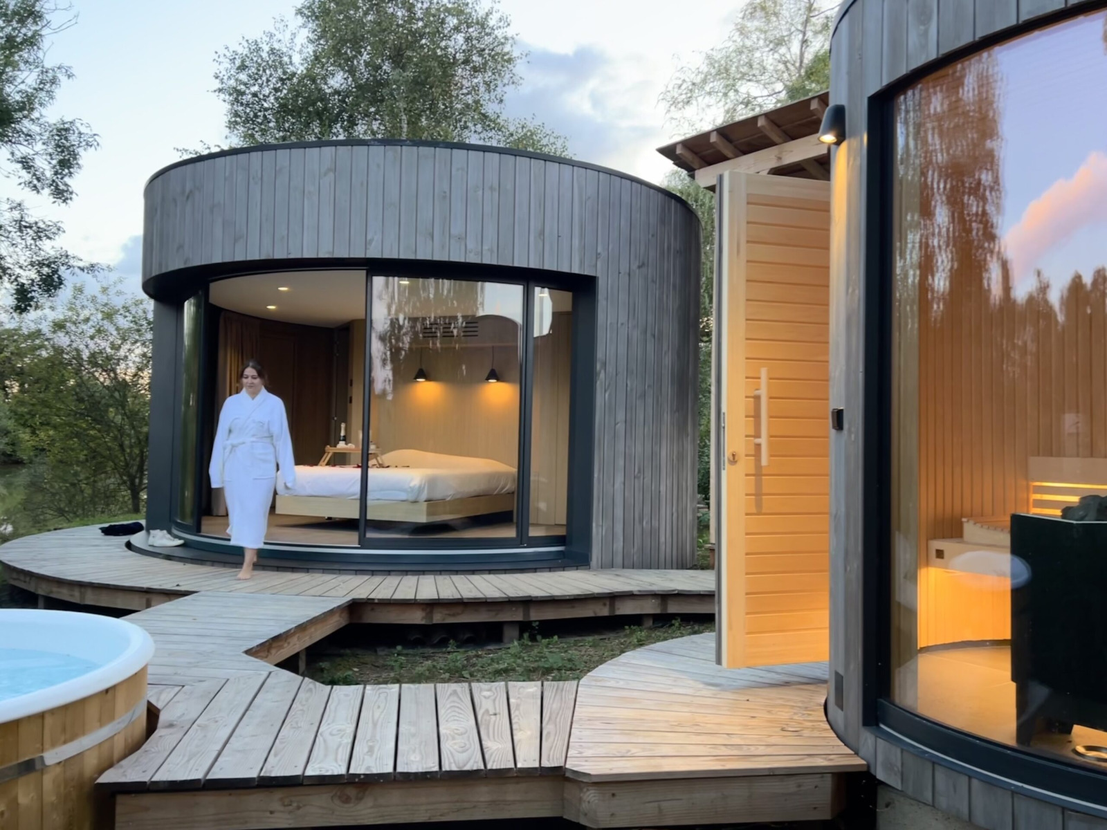 Vrouw in badjas loopt buiten een moderne ronde houten cabin met grote ramen bij Lumipod Spa île.
