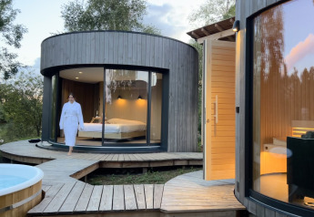 Vrouw in badjas wandelt buiten een moderne ronde houten cabin met grote ramen aan Lumipod Spa île.