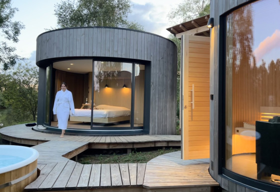 Vrouw in badjas wandelt buiten een moderne ronde houten cabin met grote ramen aan Lumipod Spa île.