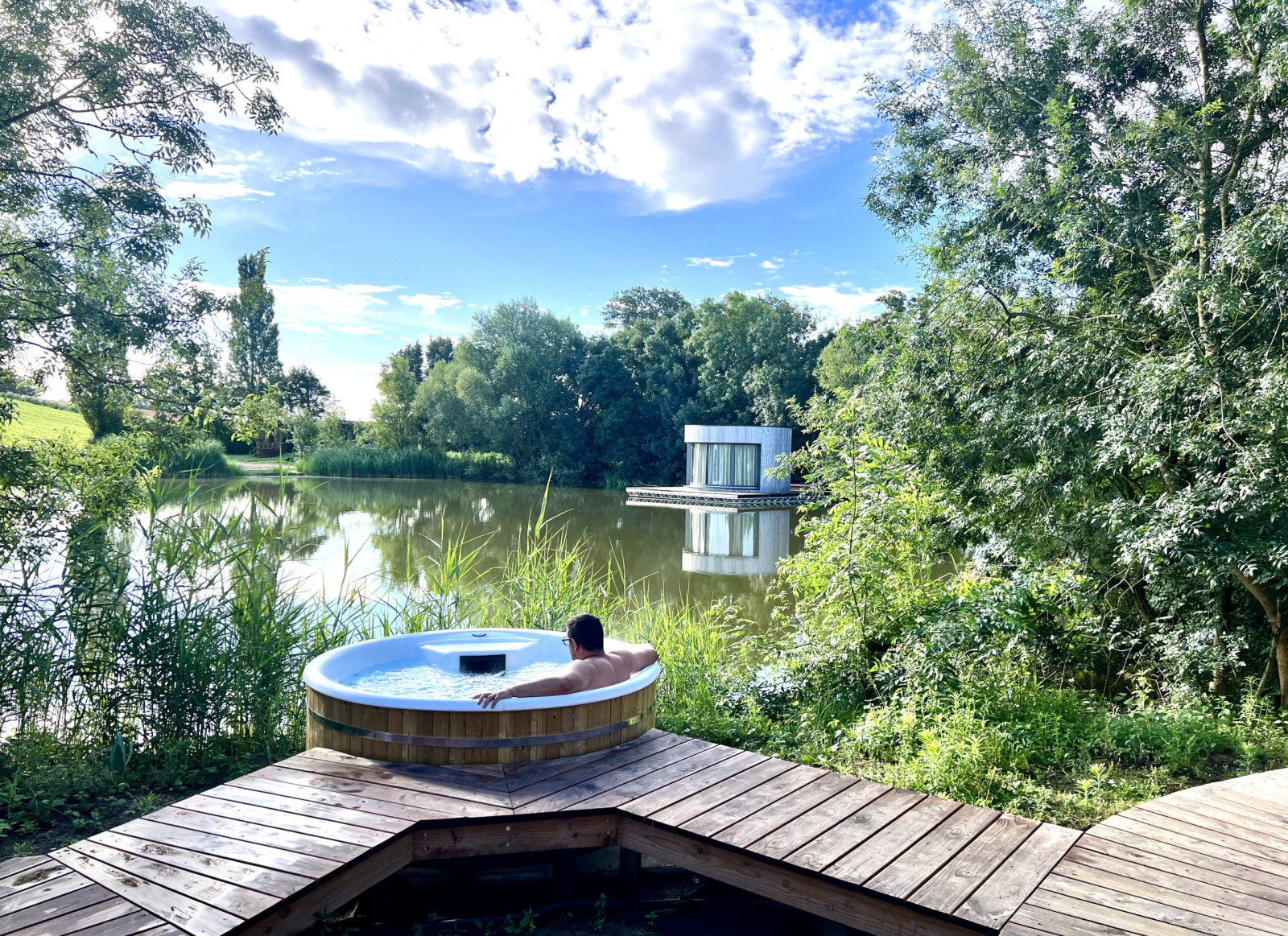 Persoon ontspant in een buitenjacuzzi met uitzicht op een meer en moderne cabine in het groene Frankrijk.
