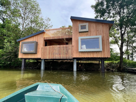 Moderne Spa Horizon Cabin op palen aan het water, gezien vanuit een boot, omringd door bomen.