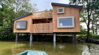 Moderna Spa Horizon Cabin su palafitte vicino all’acqua, vista da una barca, circondata da alberi.
