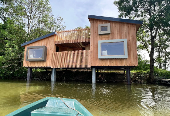 Moderne Spa Horizon Cabin op palen aan het water, gezien vanuit een boot, omringd door bomen.