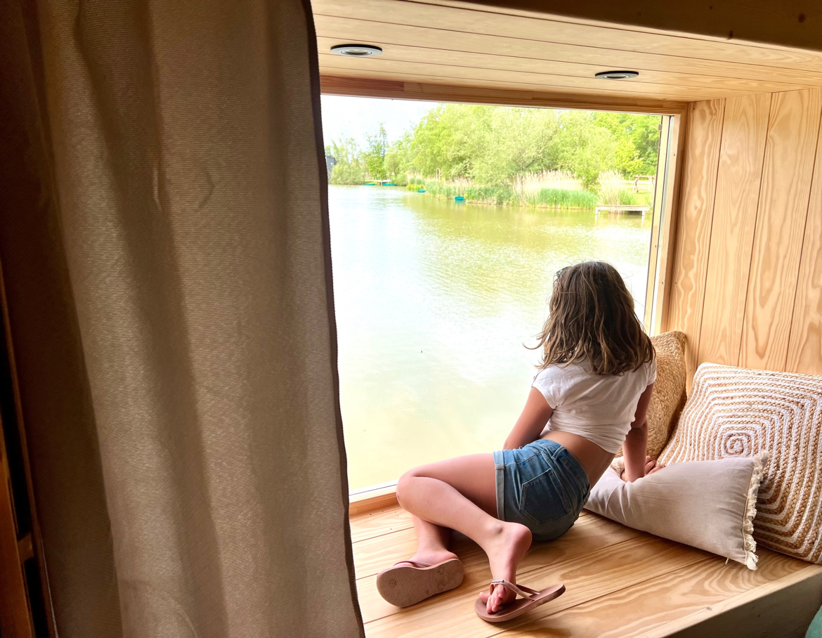 Een jonge vrouw zit in een knusse vensterbank en kijkt uit over het meer bij Spa Horizon Cabin in Domaine des Feuilles, Frankrijk.
