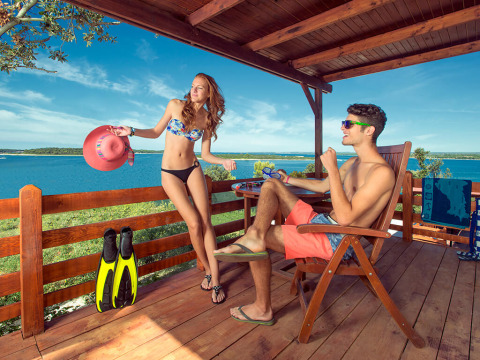 Jong stel ontspant op een houten lodge-terras met uitzicht op zee, beiden in zwemkleding en zonnebril.