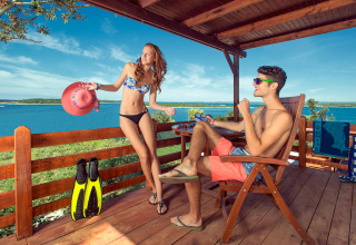 Jeune couple en maillot se détendant sur la terrasse en bois d’un lodge avec vue sur la mer en été.