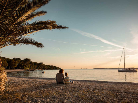 Zwei Personen sitzen am Kieselstrand bei Sonnenuntergang mit Blick auf ein Boot im Camping Brioni, Istrien, Kroatien.