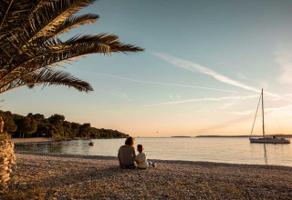 Two people sitting on a pebble beach at sunset, looking at a boat at Camping Brioni holiday park in Istria, Croatia.