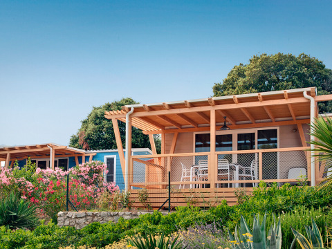 Marina Home-hut met houten veranda en tuinmeubilair, omgeven door bloemen en groen onder een blauwe lucht.