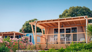 Marina Home-hut met houten terras, witte stoelen, fleurige bloemen en weelderig groen onder heldere hemel.
