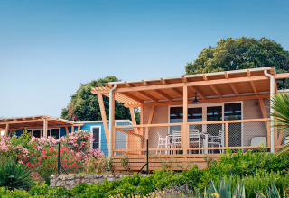 Marina Home-hut met houten veranda en tuinmeubilair, omgeven door bloemen en groen onder een blauwe lucht.