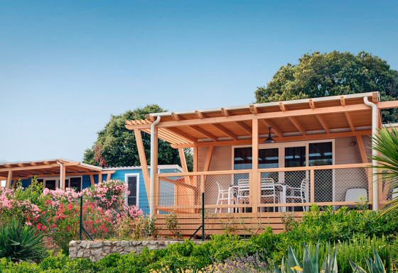 Marina Home-hut met houten veranda en tuinmeubilair, omgeven door bloemen en groen onder een blauwe lucht.