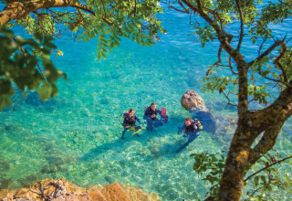 Three scuba divers in clear turquoise water under green trees, near steps and rocks in Labin, Istria, Croatia.