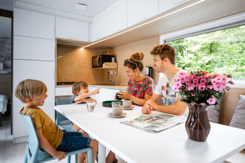 Gezin geniet van ontbijt aan tafel in moderne caravan, met bloemenvaas en buitenzicht in vakantiepark.
