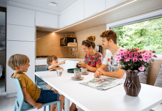 Gezin geniet van ontbijt aan tafel in moderne caravan, met bloemenvaas en buitenzicht in vakantiepark.