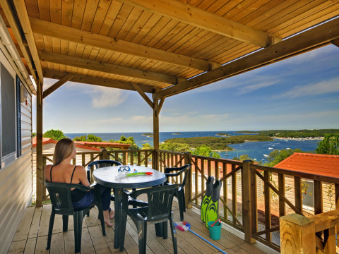 Femme assise sur la terrasse de Orsera Home au Camping Orsera, Croatie, avec vue sur la mer et les bateaux.