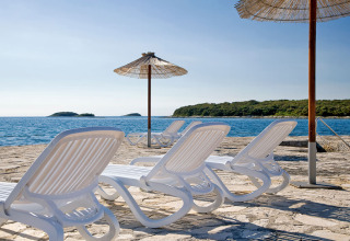 Lettini e ombrelloni al Camping Orsera in Istria, Croazia, con vista sul mare e su un’isola verde.