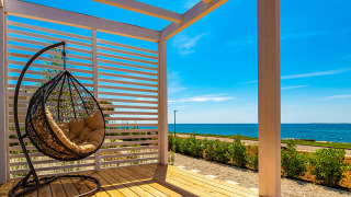 Terraza soleada de chalet con silla colgante, suelo de madera y vista al mar bajo un cielo azul.