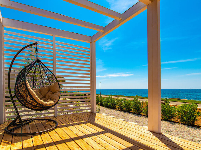 Terrazza soleggiata di uno chalet con sedia sospesa, pavimento in legno e vista mare sotto cielo azzurro.