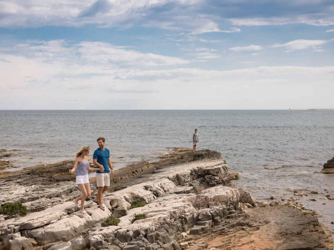 A couple walks on rocky shore at Camping Arena Grand Kažela, Istria, Croatia, with the sea in the background.