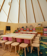 Indendørs børneområde i et tipitelt med farverige stole, borde, legetøj og opbevaringskasser i Grand Kažela.