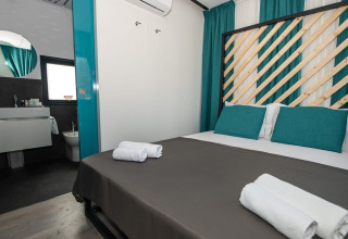 Chaletkamer met tweepersoonsbed, turquoise gordijnen en kussens, badkamergedeelte en propere handdoeken.