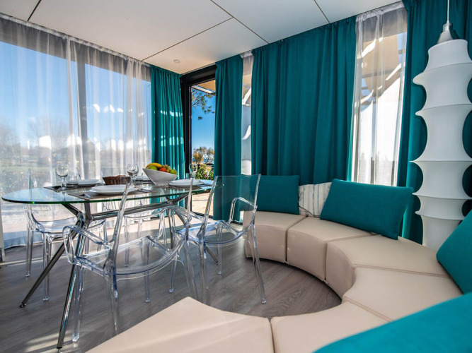 Moderne chaletwoonkamer met ronde zetel, glazen tafel, doorzichtige stoelen en turquoise gordijnen.